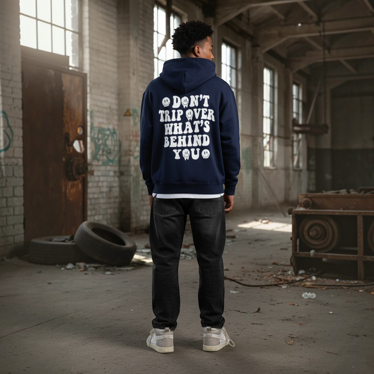 BY JAXXIE donkerblauwe oversized hoodie met witte tekstprint op de rug, gedragen door een man in een industriële hal.