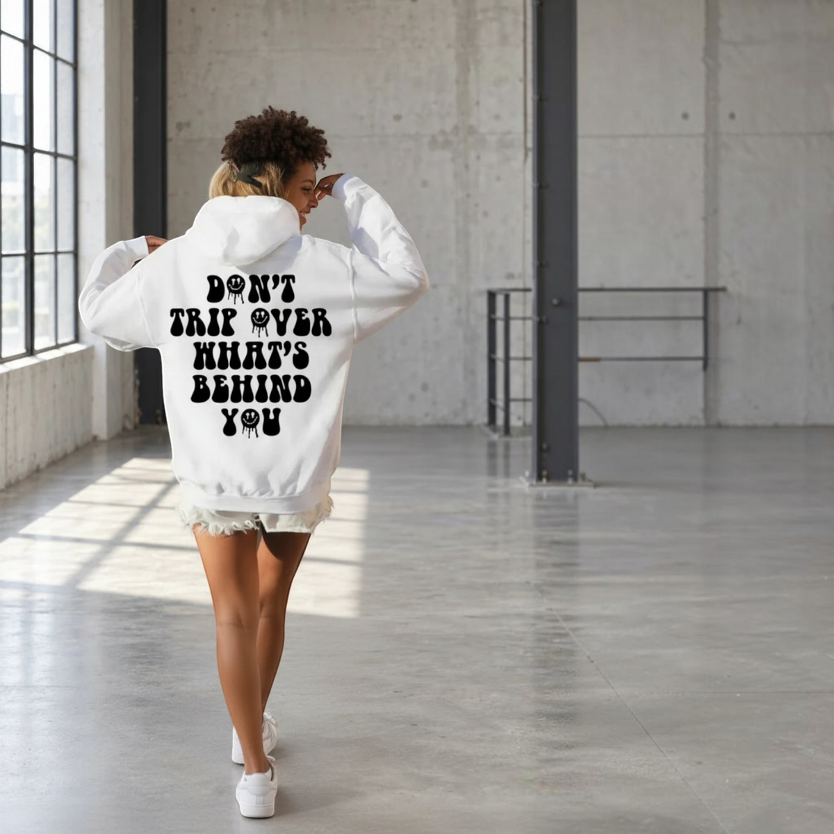 BY JAXXIE witte oversized hoodie met zwarte Don't Trip backprint gedragen door een vrouw in een industriële ruimte